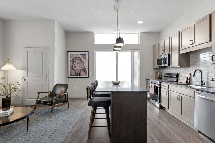 Modern open-concept apartment kitchen at Harlow in Chanhassen, MN, featuring a spacious island with bar seating, light wood cabinetry, stainless steel appliances, subway tile backsplash, pendant lighting, and an adjoining living area.